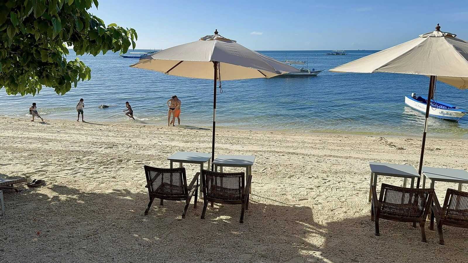 Bounty Beach Malapascua Cebu sandy beach scene with umbrellas, beach chairs, kids playing in shallow turquoise water, and bangka boats moored offshore